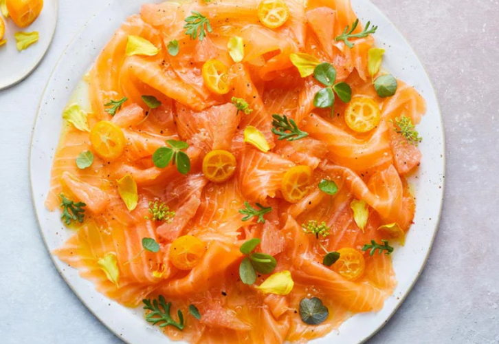 Smoked salmon carpaccio with citrus fruits, kumquat slices, finger lime pearls, microgreens, and edible flowers on a white plate