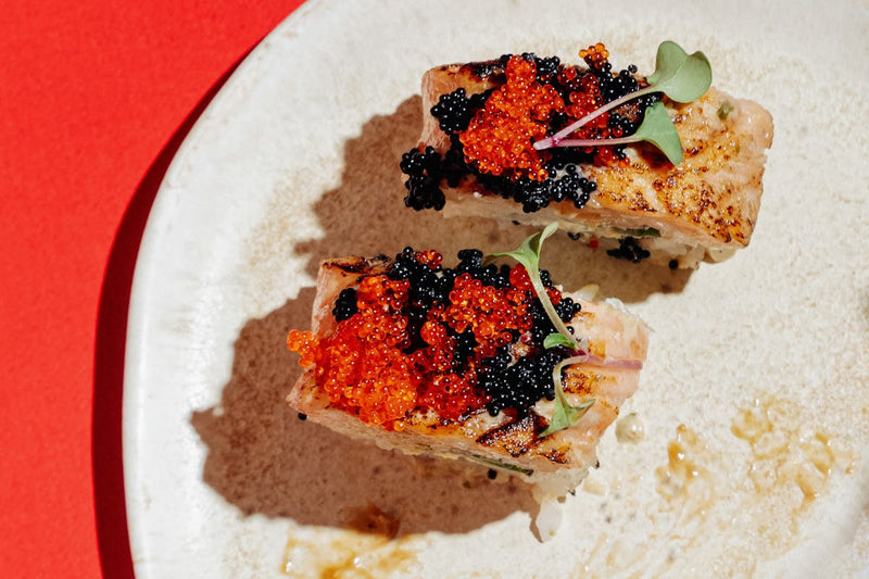 Overhead flat-lay of caviar tin with mother-of-pearl spoon, blinis, and scrambled eggs on marble surface for elegant caviar recipes table setting