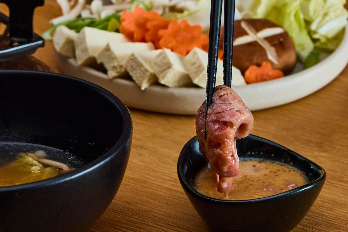 British Wagyu Beef being dipped in sauce with chopsticks alongside a plate of sliced vegetables and tofu for hot pot
