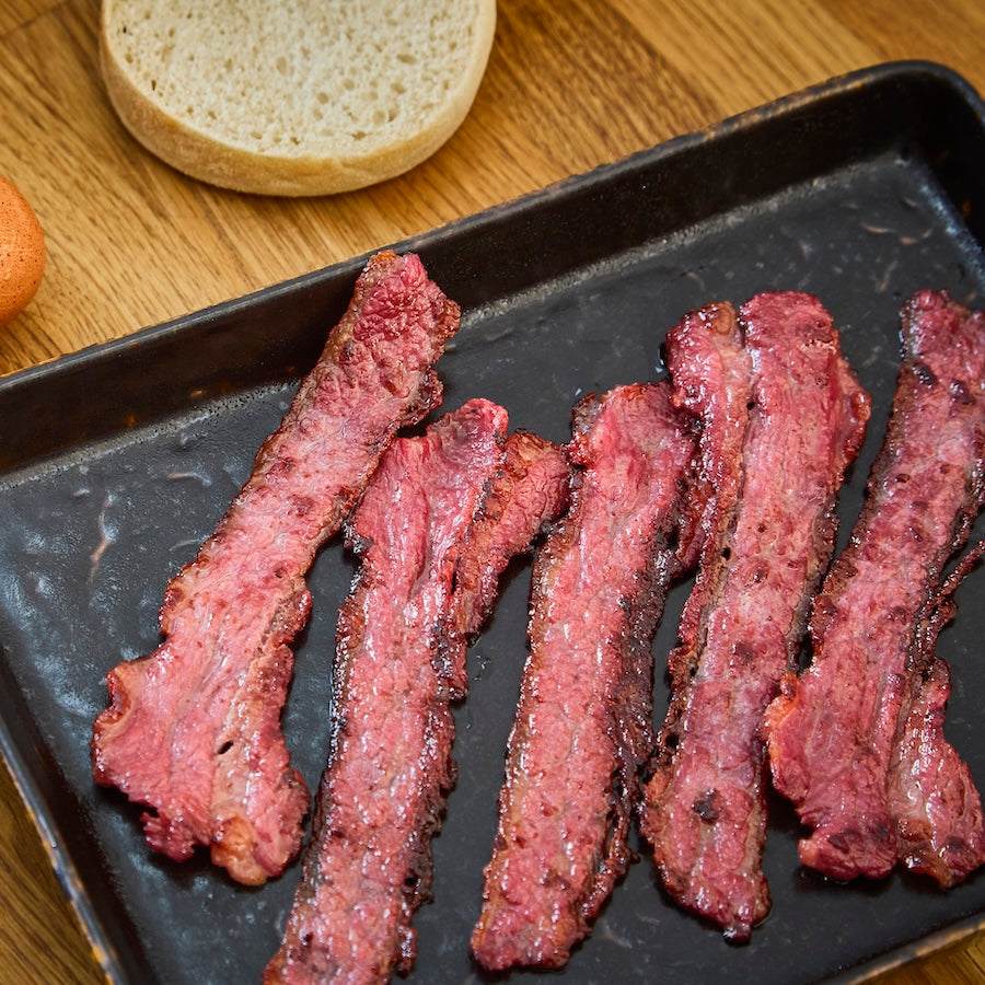 Wagyu British Beef Bacon cooked to crispy, rich reddish-brown strips on a black tray beside a bread roll slice