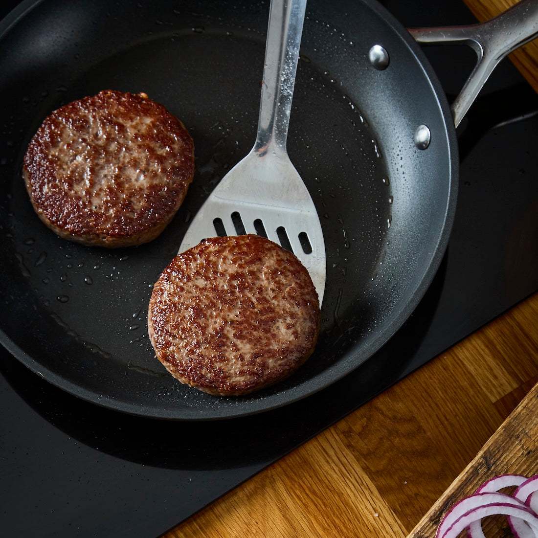 Two browned Wagyu British Beef Burgers cooking in a black frying pan with a metal spatula on a black stovetop