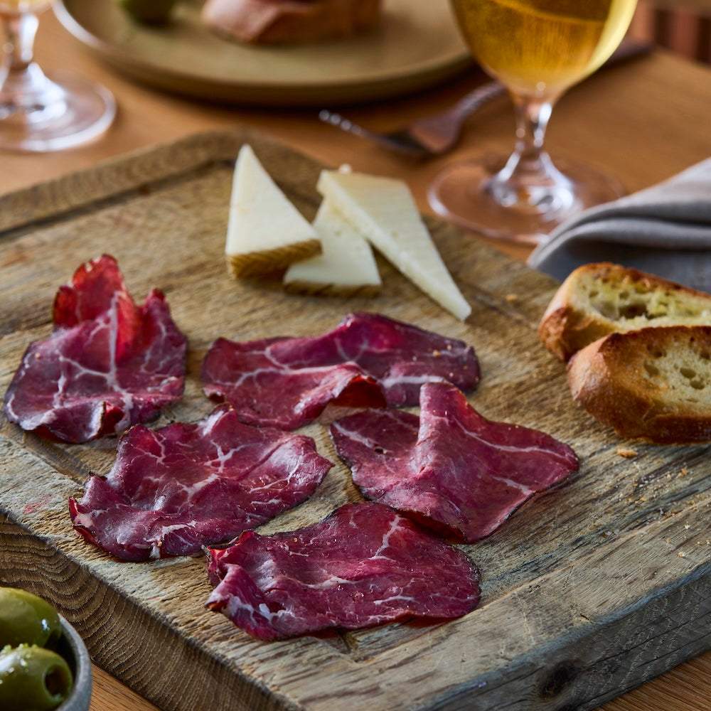 Wagyu British Beef sliced bresaola with deep red marbled texture arranged on wooden board with cheese wedges and toasted bread