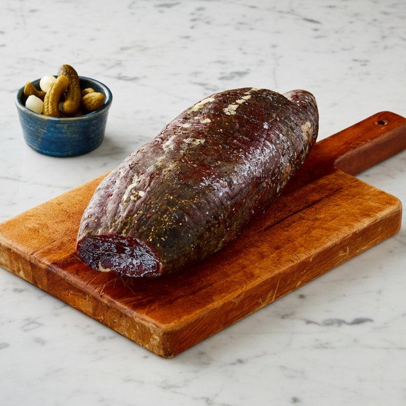 Wagyu British Beef whole bresaola with dark cured exterior on wooden cutting board with small bowl of pickles